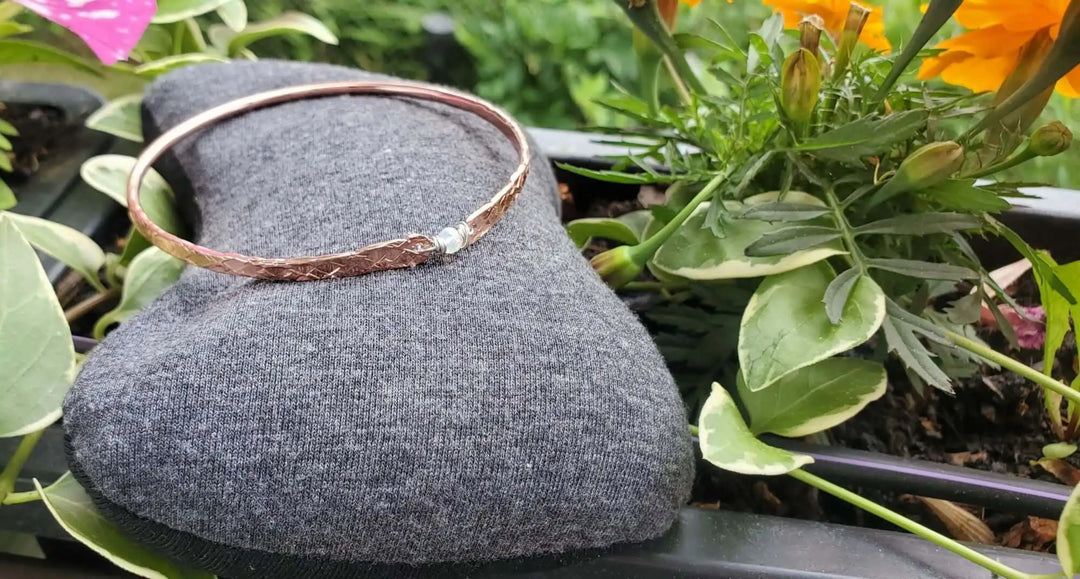 Versions of Best Selling Featured Copper Brides Mag Bangle on a grey display amidst vibrant flowers.