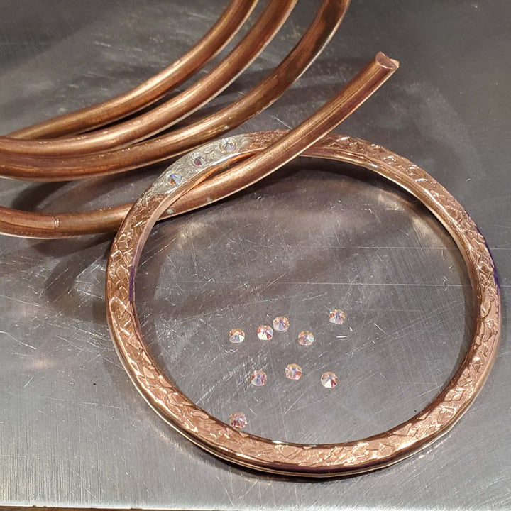 A copper ring band with a textured hammered finish and a matching coiled copper tube, displayed on a metallic workbench surface.
