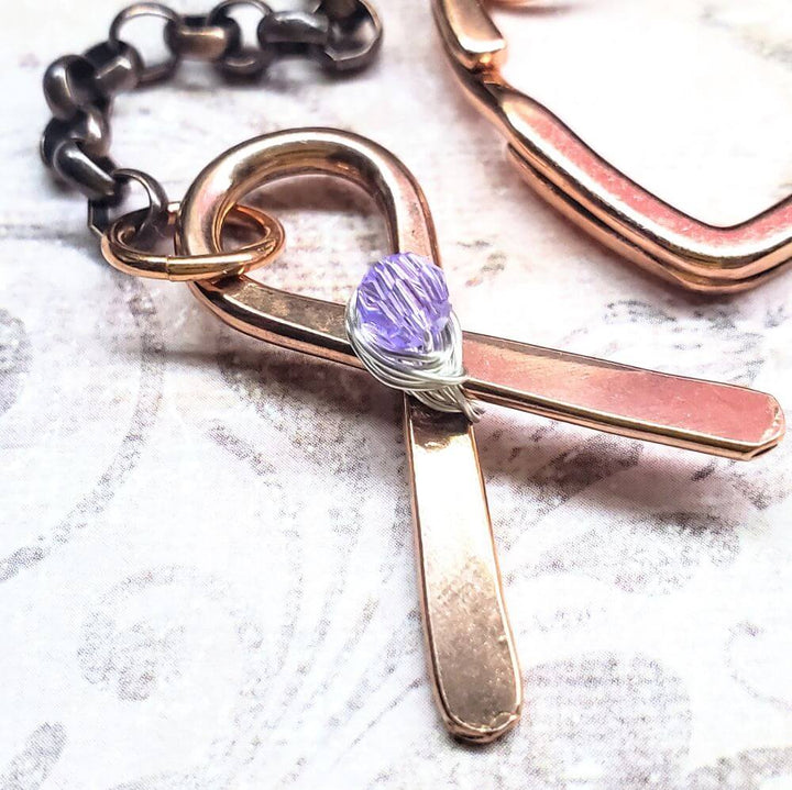 Rose gold cross keychain with purple gemstone on a textured surface