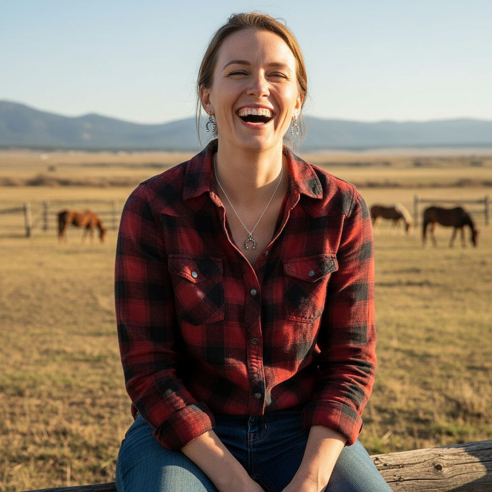 Woman wearing handmade sterling silver horseshoe jewelry, smiling outdoors with horses in the background.