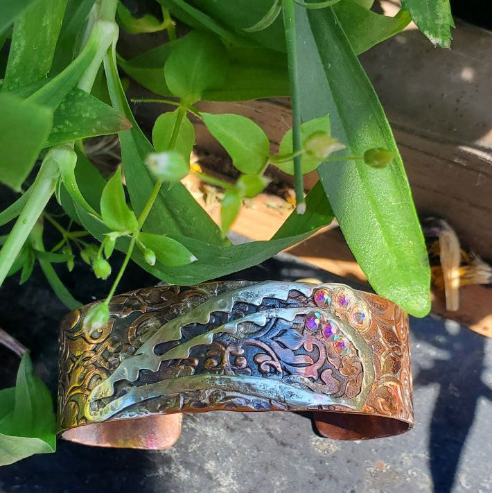 A hammered metal cuff bracelet with a warm copper-brown base and iridescent blue, purple, and green shifts, resting on a dark surface with green plants in the background.