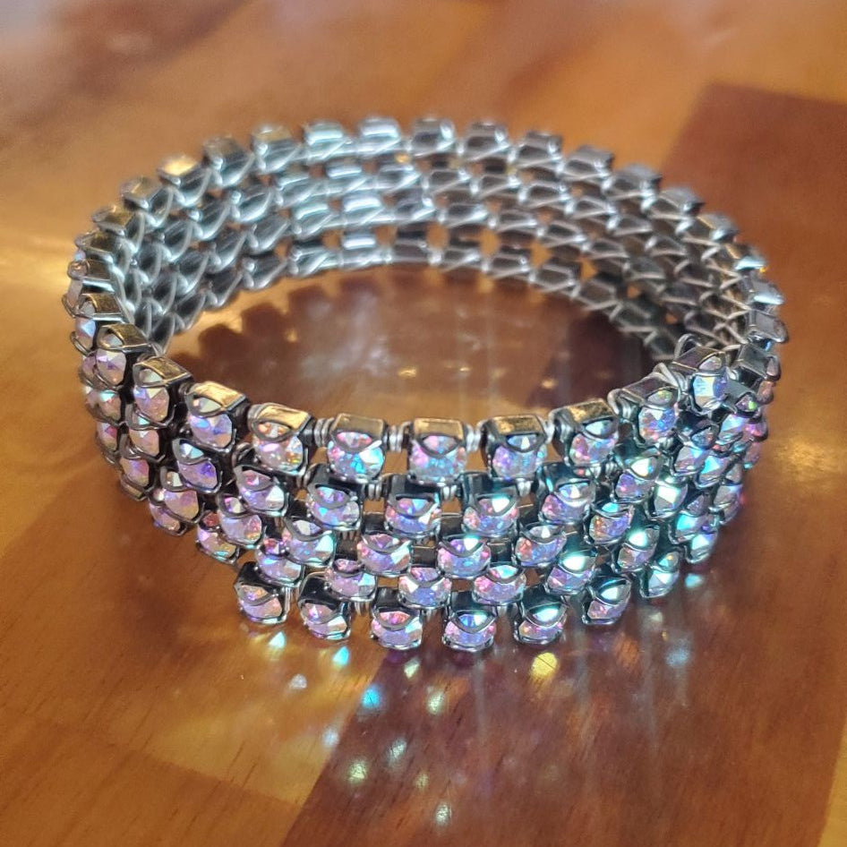 A wide, multi-strand rhinestone bracelet with silver-toned metal and iridescent stones, resting on a polished wooden table.