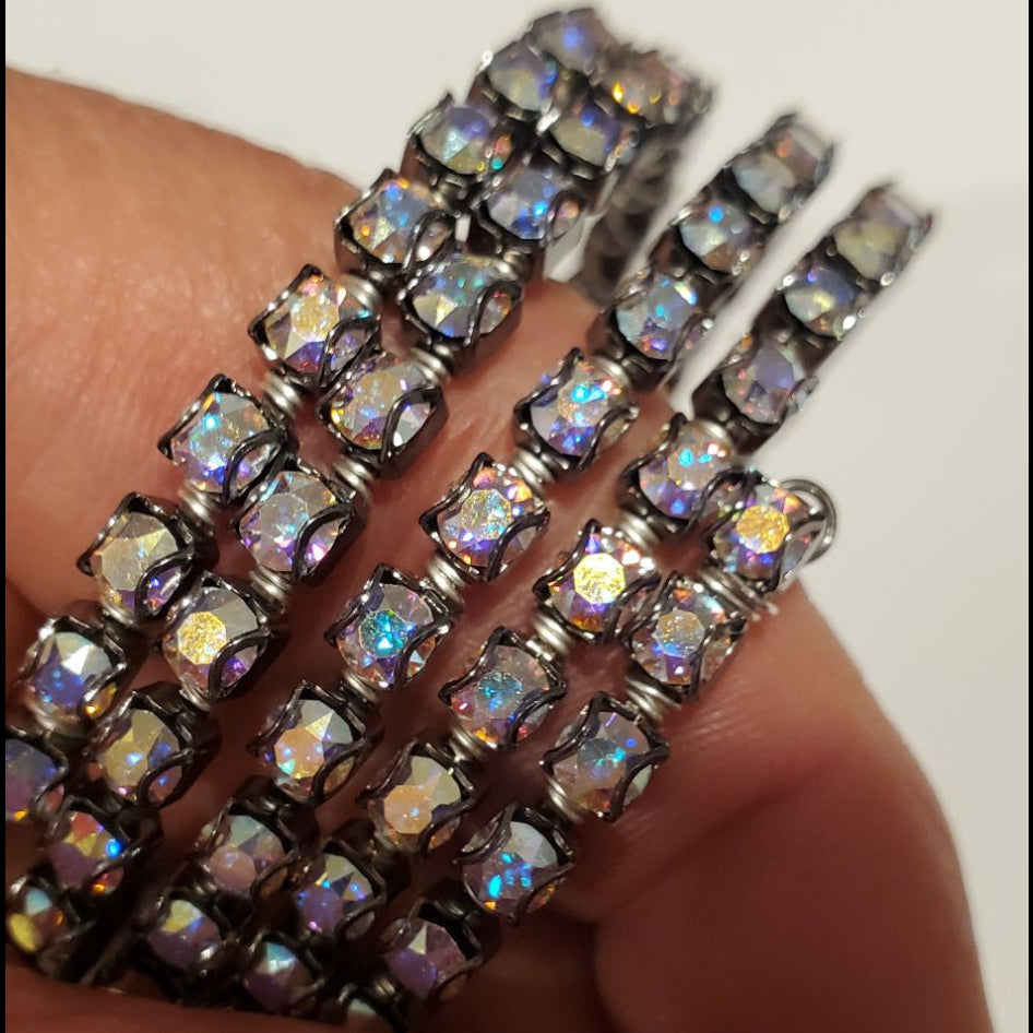 A close-up of a hand holding a stack of silver-toned bracelets adorned with iridescent, rainbow-reflecting rhinestones against a plain light background.