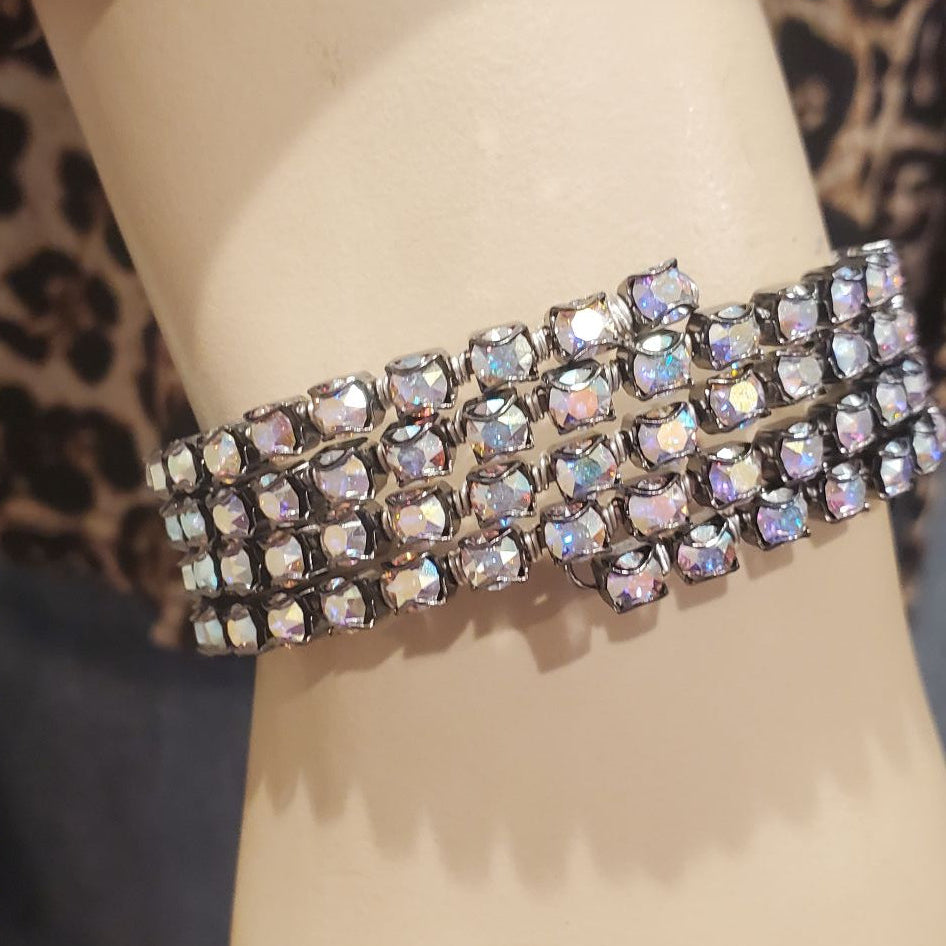 A wide, multi-row bracelet covered in iridescent rhinestones, displayed on a beige mannequin arm with a leopard-print fabric and blue jeans in the background.