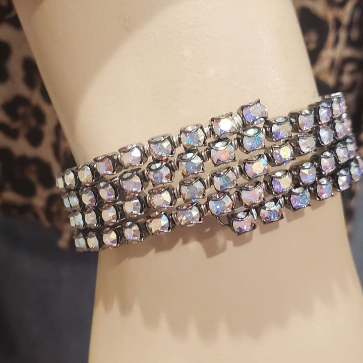A wide, multi-row bracelet covered in iridescent rhinestones, displayed on a beige mannequin arm with a leopard-print fabric and blue jeans in the background.