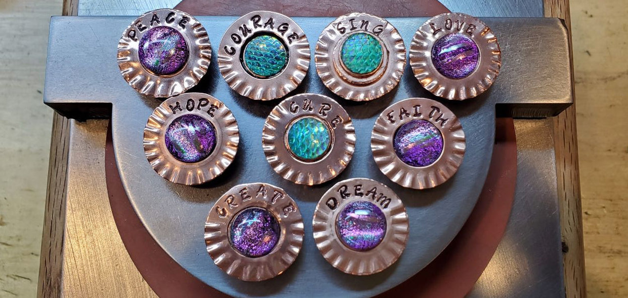 A close-up of a handmade jewelry-making setup showing multiple copper coin bezels with colorful glass inlays (purple, green, blue) arranged on a gray metal stand over a wooden workbench.