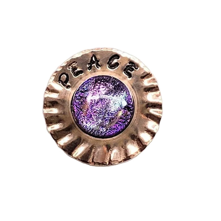 A small round jewelry piece with a purple center and a bronze-toned sunburst border, shown on a white surface with a hand partially visible in the lower right corner.