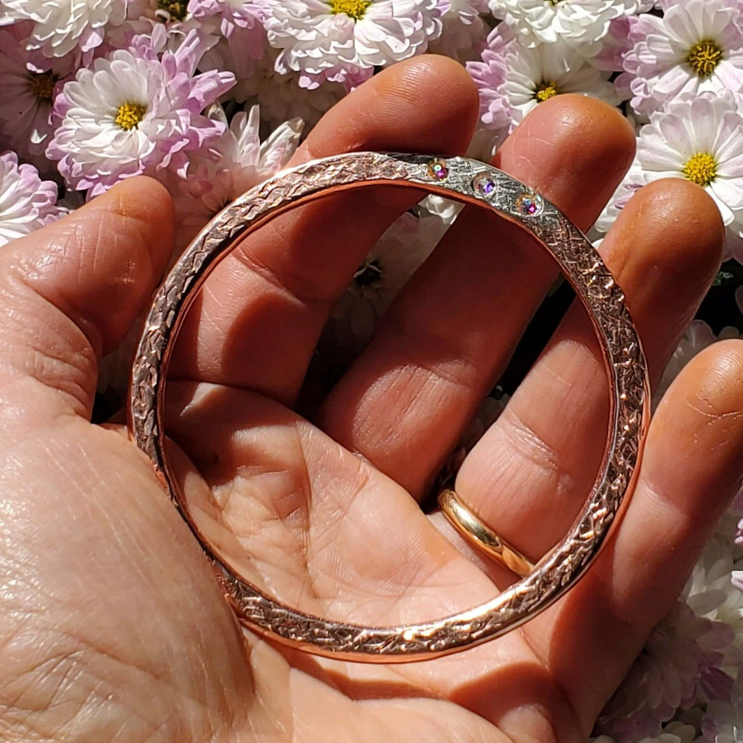 A rose gold bangle bracelet with a textured, twisted outer pattern and a smooth inner band, held in a person&