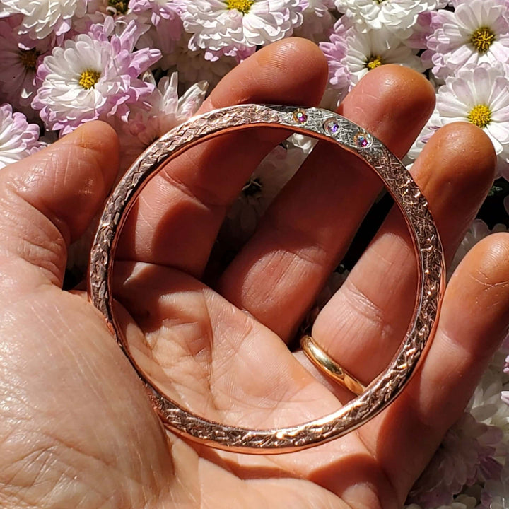 A rose gold bangle bracelet with a textured, twisted outer pattern and a smooth inner band, held in a person&