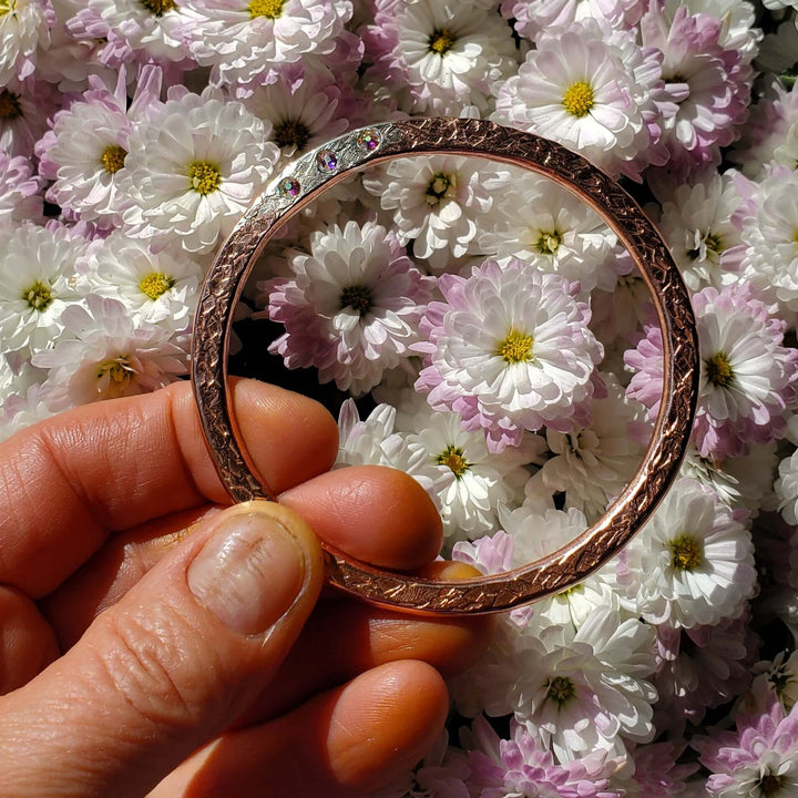 A hand holding a thin rose gold bangle bracelet in front of a dense background of white and pink chrysanthemums.