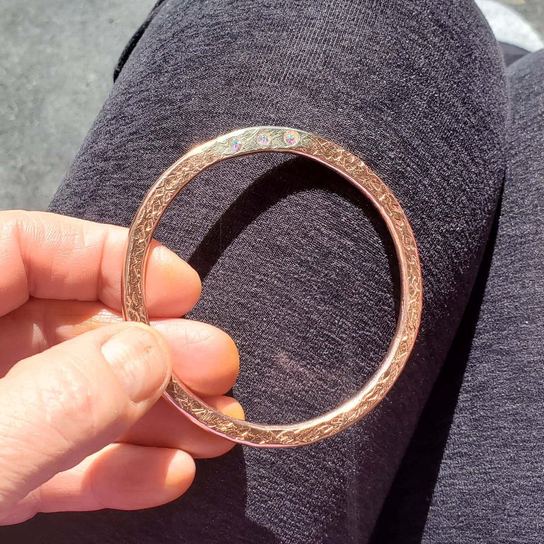 A hand holding a thick, polished rose gold bangle with three crystal inlays against a dark gray textured background.
