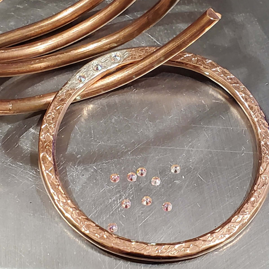 A copper bangle bracelet with a textured patterned border and a matching coiled copper wire, displayed on a brushed metal surface.