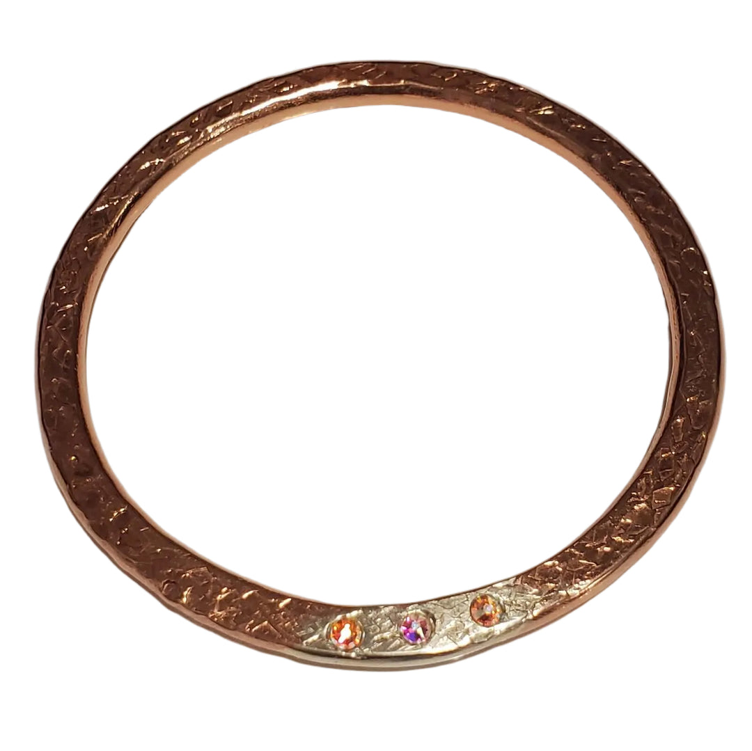 A rose gold bangle bracelet with engraved text and a small iridescent crystal accent, resting on a light surface.