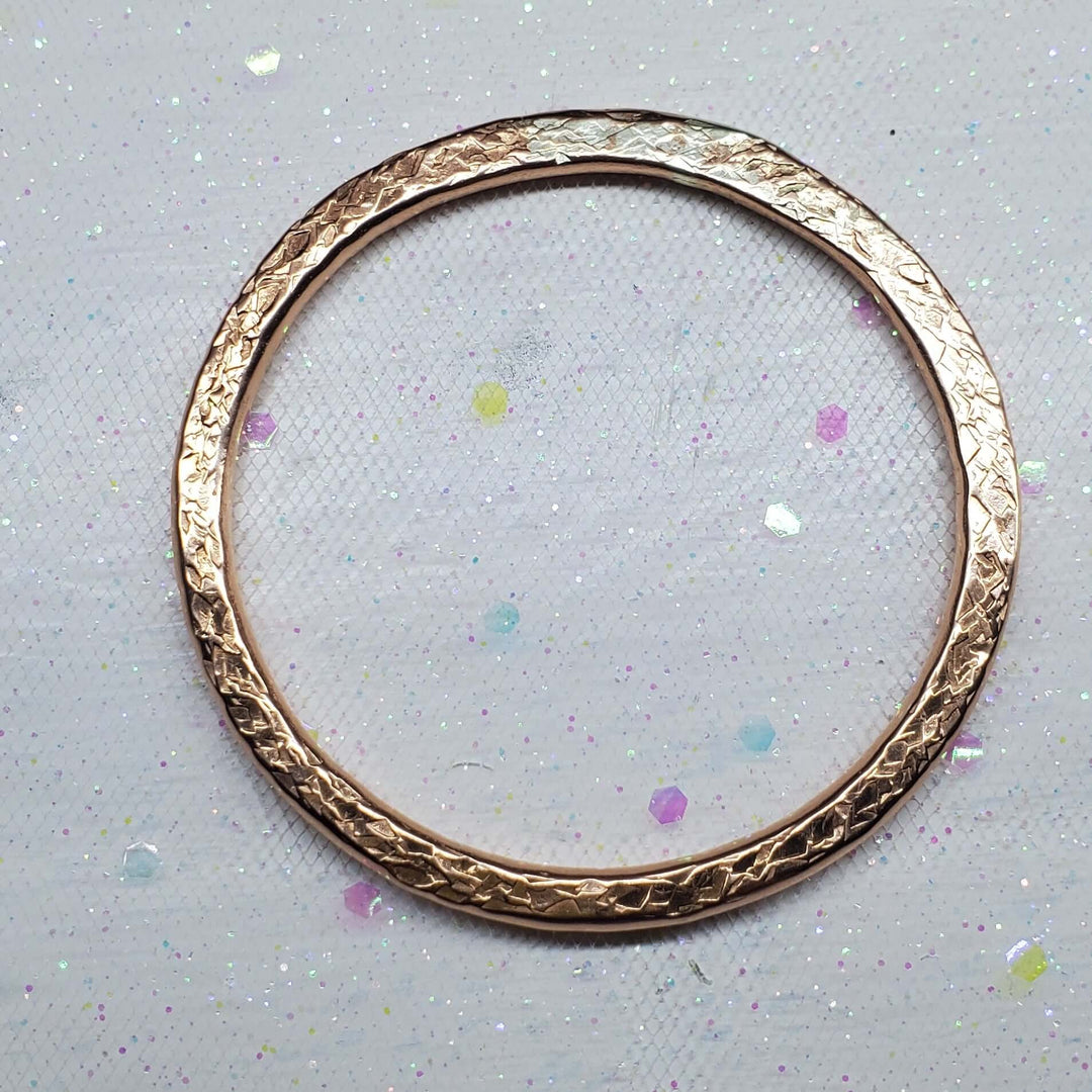 A single rose gold bangle bracelet with a textured, hammered finish, displayed on a white glittery fabric background.