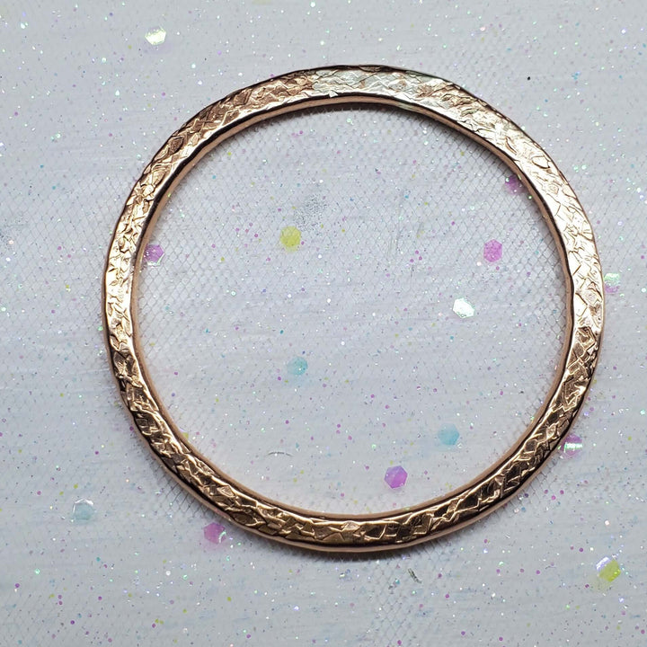 A single rose gold bangle bracelet with a textured, hammered finish, displayed on a white glittery fabric background.