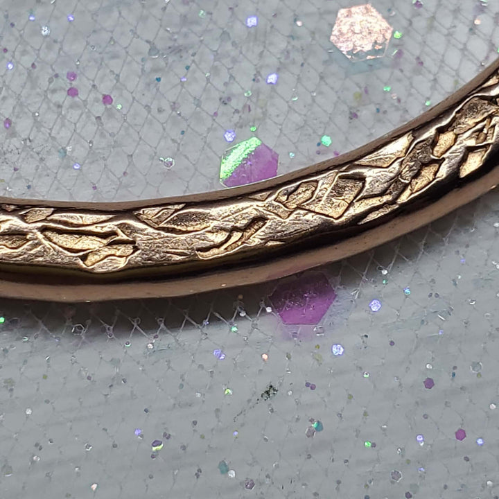 A close-up of a gold-toned metal bangle bracelet with an intricate leaf or vine pattern, resting on a textured gray glittery surface.