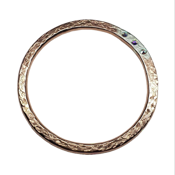 A single rose gold textured bangle bracelet with a small silver-colored stone set on the outer edge, displayed on a glittery light gray fabric background.