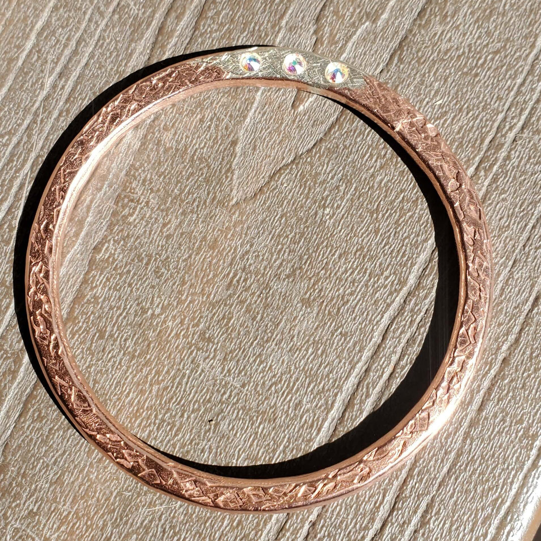 A rose gold bangle bracelet with a textured floral pattern and three small clear stones, resting on a textured gray surface.
