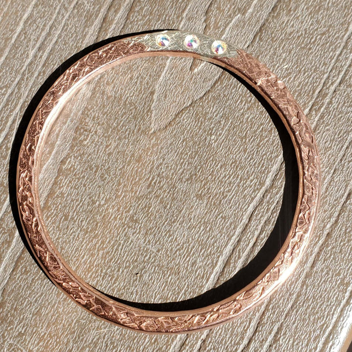 A rose gold bangle bracelet with a textured floral pattern and three small clear stones, resting on a textured gray surface.