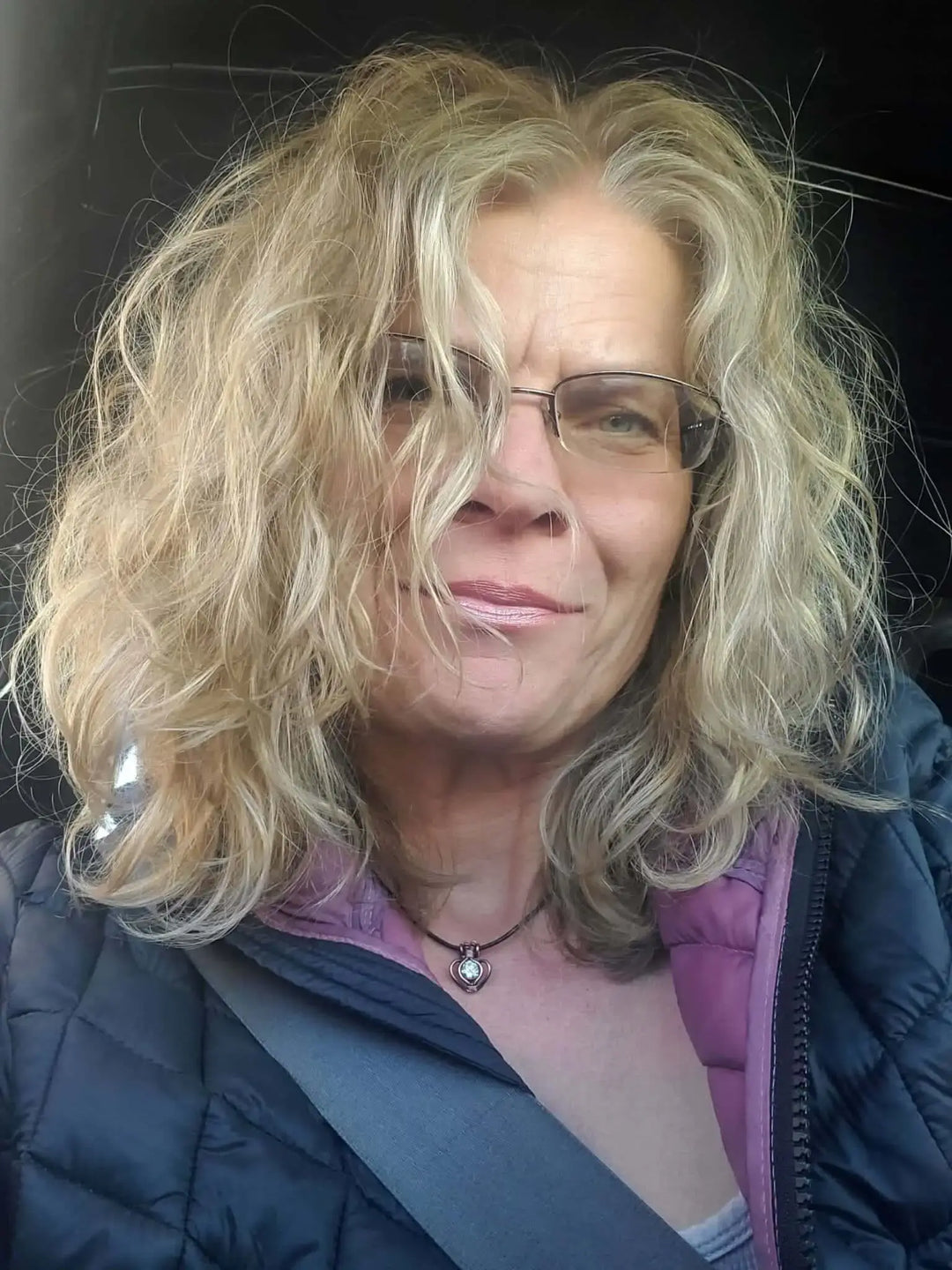 Close-up of a woman with curly hair wearing glasses and a necklace, smiling while seated in a vehicle.