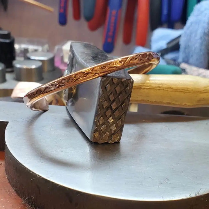 Handmade 2 ounce 6 Gauge Copper Hammered BIGGA Bangle resting on a hammer, showcasing its unique design and craftsmanship.