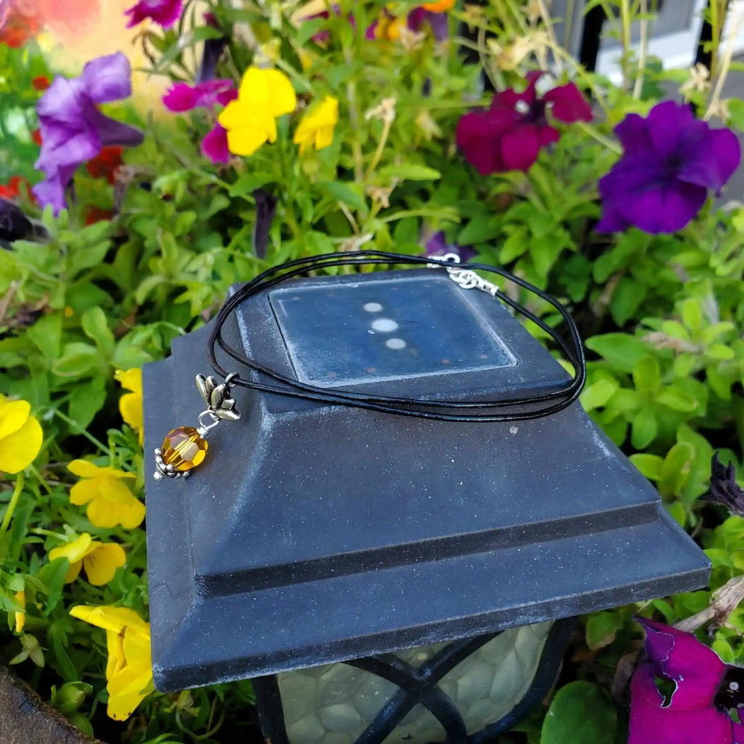 Handmade silver crystal pineapple leather necklace displayed on a lantern surrounded by colorful flowers.