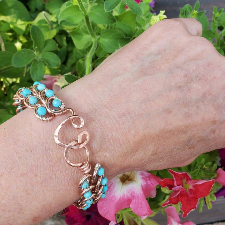 Wire Wrapped Turquoise Copper Infinity Heart Closure Bangle worn on wrist surrounded by colorful flowers.