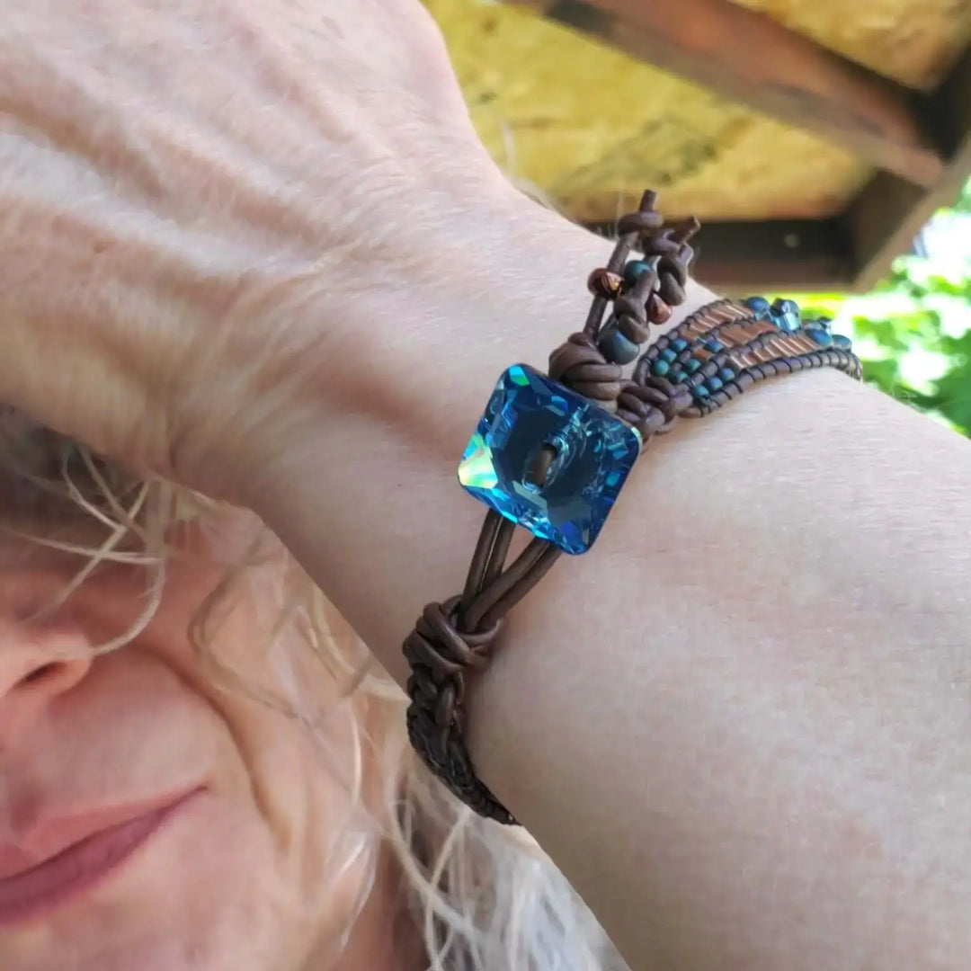 Intricate Bead Woven Swarovski Crystal Button Glam Bracelet displayed on a wrist with a blue crystal centerpiece and leather wrap.