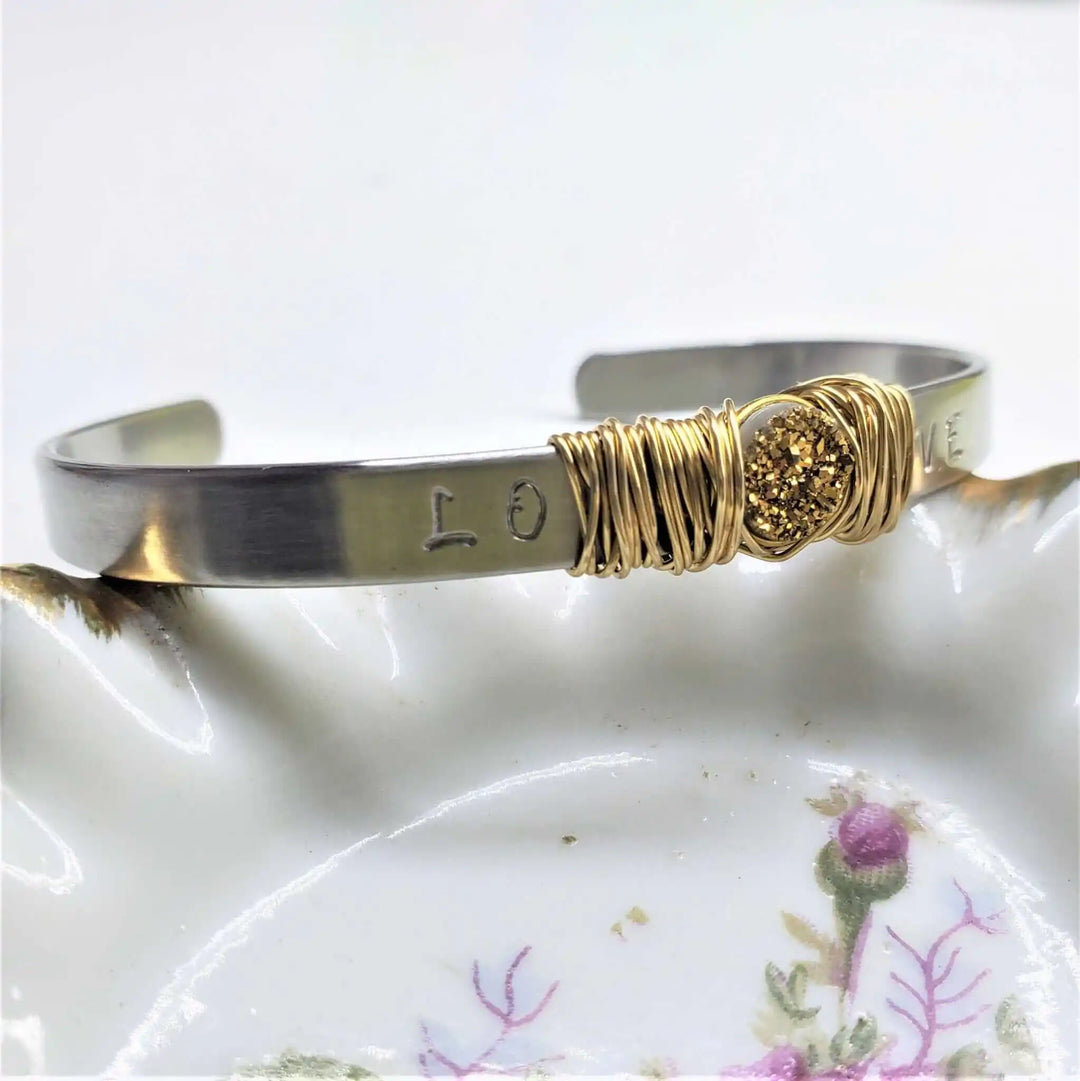Stainless Steel LOVE Stamped Wire Wrapped Gold Druzy Cuff displayed on a floral plate, highlighting its unique design.