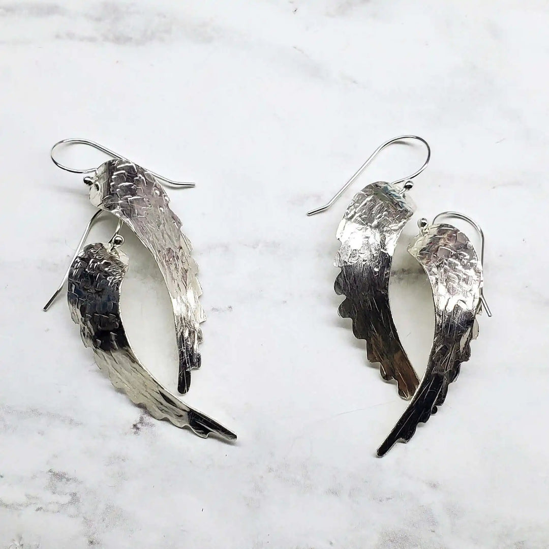 Silver Hammered Sculpted Angel Wing Earrings displayed on a marble surface, showcasing their intricate hammered texture.