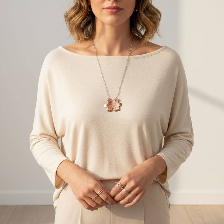 Woman wearing a beige long-sleeve top with a necklace and rings against a neutral background