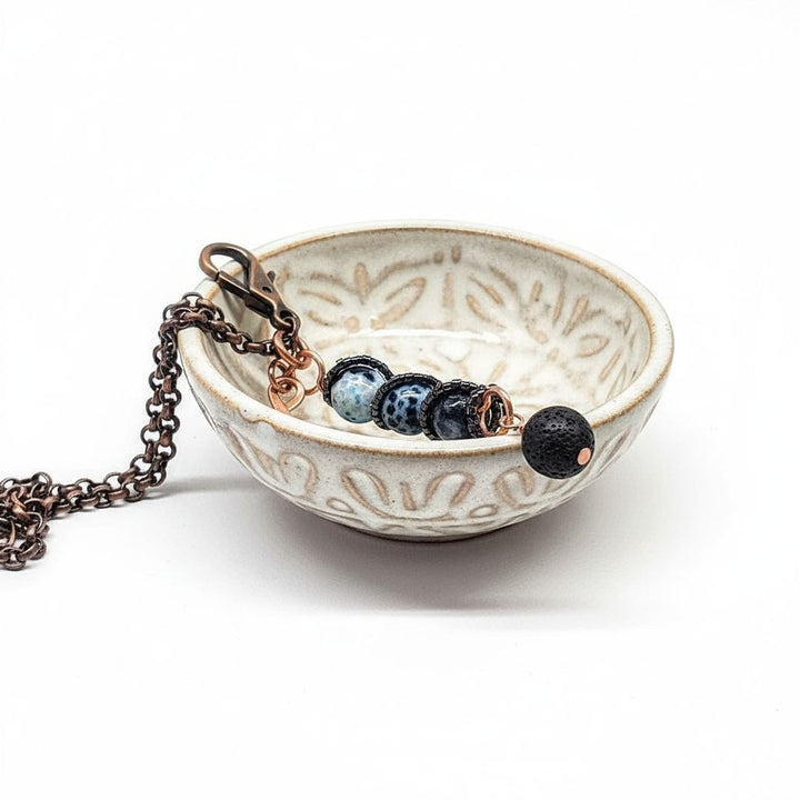 Child Abuse Awareness Agate Necklace With Lava Rock Bead displayed in a decorative bowl, featuring blue agate and lava rock beads.