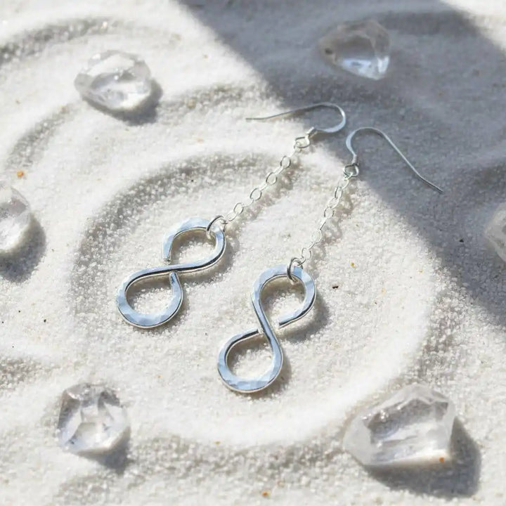 Sterling Silver Hammer Patterned Infinity Earrings displayed on sandy background with crystals.