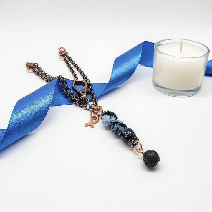 Child Abuse Awareness Agate Necklace With Lava Rock Bead featuring blue agate and lava rock beads, displayed with a blue ribbon and candle.