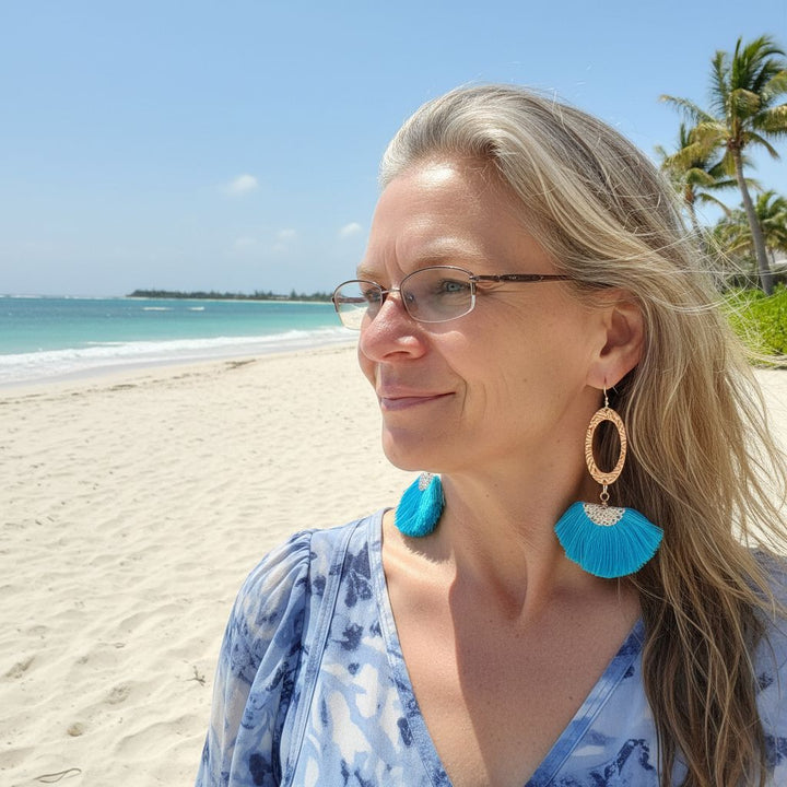 Long Copper Embossed Aqua Brushed Silk Fan Tassel Earrings worn by a woman on the beach with the ocean in the background.